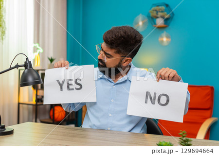Indian man holds YES and NO signs uncertain choice seeking answer confused decision making dilemma 129694664