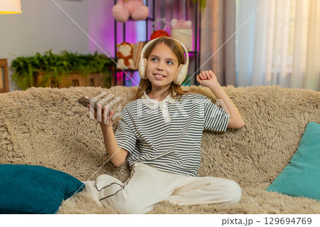 Child girl kid joyfully listens music on smartphone with wireless headphones while dancing on sofa Child girl kid joyfully listens music on smartphone with wireless headphones while dancing on sofa 129694769