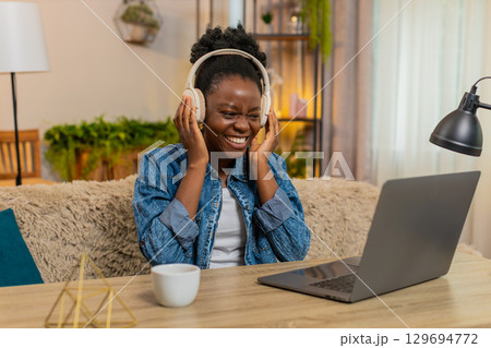 African American young woman dancing while listening to music in wireless headphones at home on sofa African American young woman dancing while listening to music in wireless headphones at home on sofa 129694772
