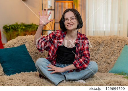 Young woman smiling and waving hand at home, greeting someone with friendly and warm expression Young woman smiling and waving hand at home, greeting someone with friendly and warm expression 129694790