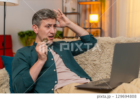 Young man holds bitcoin coin and reacts with stress after seeing crypto market drop on laptop screen 129694808