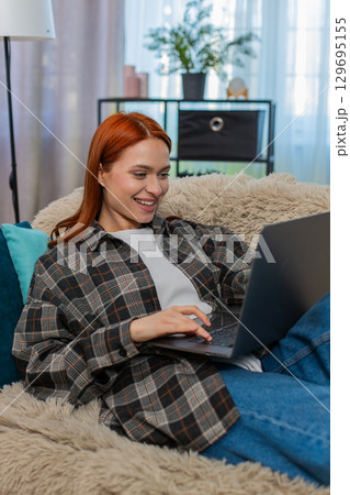 Woman sitting on sofa shopping online with laptop choosing dress touching chin in thoughtful pose Woman sitting on sofa shopping online with laptop choosing dress touching chin in thoughtful pose 129695155