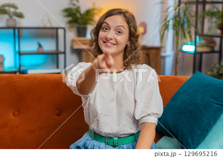 Young woman smiling and pointing to camera, choosing inviting, select sitting on sofa couch at home 129695216
