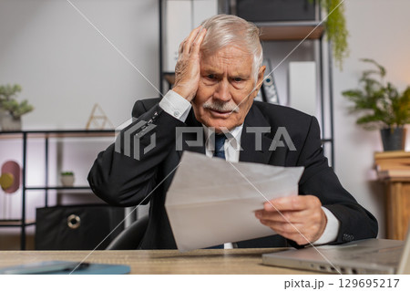 Senior businessman opening envelope reading bad news in letter feels upset sitting at office desk 129695217