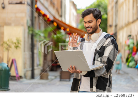 Young Indian man freelancer sitting on street, looking at laptop camera, making video call outdoors Young Indian man freelancer sitting on street, looking at laptop camera, making video call outdoors 129695237