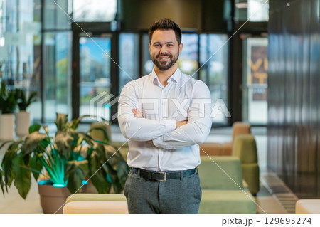 Portrait of happy young businessman with arms crossed looking at camera in modern office hotel lobby 129695274