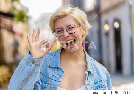 Portrait of attractive young millennial woman looking at camera showing hand OK sign on city street 129695531