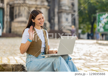 Happy young woman using laptop having video call chat conversation while sitting in city street 129695666