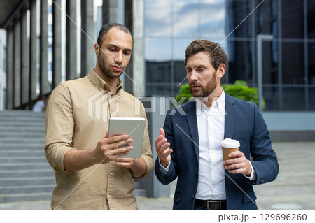 Two individuals engage in professional discussion looking at a tablet outdoors. The environment features modern architecture, indicating a corporate setting that highlights business collaboration Two individuals engage in professional discussion looking at a tablet outdoors. The environment features modern architecture, indicating a corporate setting that highlights business collaboration 129696260