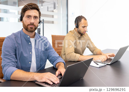 Two office professionals using laptops, wearing headsets, and engaged in a focused environment. They collaborate in an office setting, symbolizing communication, teamwork, and technology-driven Two office professionals using laptops, wearing headsets, and engaged in a focused environment. They collaborate in an office setting, symbolizing communication, teamwork, and technology-driven 129696291