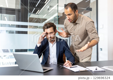 Two colleagues in an office argue over work-related matters while using a laptop. 129696324