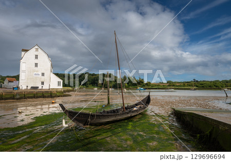 Old wooden boat with mast sitting on mud. Old tide mill behind. View of the quay at low tide in the River Deben at the historic town of Woodbridge in Suffolk, UK. 129696694
