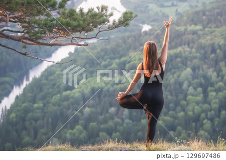Woman is performing yoga on a mountaintop during sunrise, surrounded by a serene landscape. tands tree pose, with her right arm raised in a mudra. Below her, a river winds through a densely forested Woman is performing yoga on a mountaintop during sunrise, surrounded by a serene landscape. tands tree pose, with her right arm raised in a mudra. Below her, a river winds through a densely forested 129697486