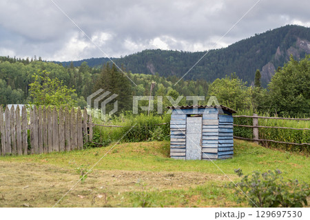 Rustic old outdoor toilet blue house on the background of beautiful nature and mountains. Small rustic shed stands against a backdrop of green forests and mountains under a cloudy sky 129697530