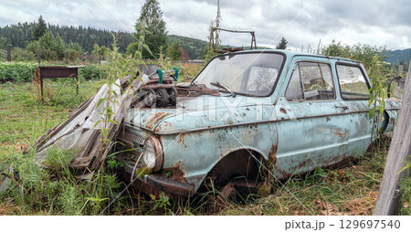 An old Soviet abandoned car Zaz Zaporozhets. Old rusted car abandoned in a field surrounded by overgrown grass and trees under a cloudy sky. An old Soviet abandoned car Zaz Zaporozhets. Old rusted car abandoned in a field surrounded by overgrown grass and trees under a cloudy sky. 129697540