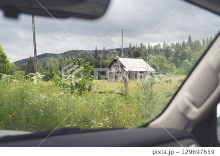 Old wooden cabin nestled in the lush green countryside viewed from a vehicle on a cloudy day in the forested mountains. Road trip to remote locations in Siberia, view from the car. 129697659