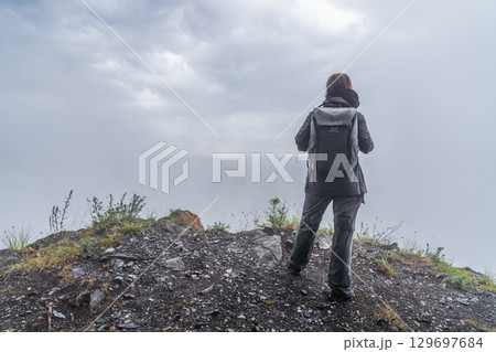 A person stands at the edge of a rocky hillside, gazing into the distance as low clouds create a misty atmosphere. The early morning light pierces through the gray sky, illuminating the damp terrain. A person stands at the edge of a rocky hillside, gazing into the distance as low clouds create a misty atmosphere. The early morning light pierces through the gray sky, illuminating the damp terrain. 129697684