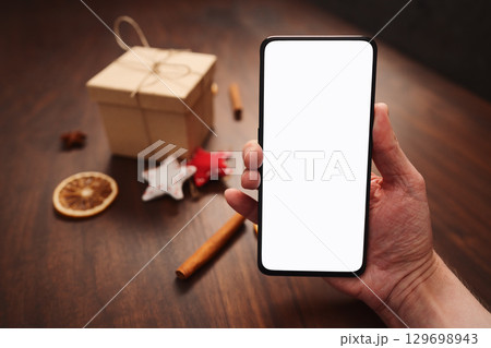 Man hand hold smarthone with white screen with christmas decorations on background Man hand hold smarthone with white screen with christmas decorations on background 129698943