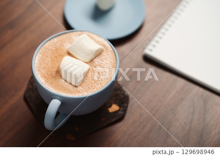 Hot chocolate in a blue cup with marshmallow and notepad on walnut table 129698946