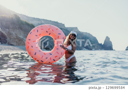 Woman, donut, ocean. Happy young woman holds inflatable donut in the sea near a rocky beach on summer vacation Woman, donut, ocean. Happy young woman holds inflatable donut in the sea near a rocky beach on summer vacation 129699584