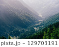Summer view into a valley in the Austrian Alps. Sunlight shines over the landscape, with forested mountains on the sides and a winding road and river at the bottom of the valley. 129703191