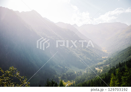 Summer view into a valley in the Austrian Alps. Sunlight shines over the landscape, with forested mountains on the sides and a winding road and river at the bottom of the valley. 129703192