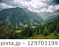 Summer view into a valley in the Austrian Alps. Sunlight shines over the landscape, with forested mountains on the sides and a winding road and river at the bottom of the valley. 129703199