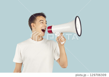 Young man or teenage boy makes loud announcement through megaphone isolated on blue background Young man or teenage boy makes loud announcement through megaphone isolated on blue background 129706309