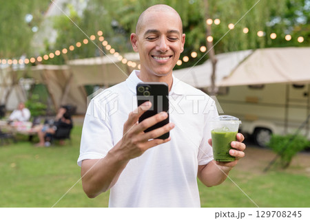 Bald man at campsite with camper van having healthy green detox juice drink and using phone 129708245