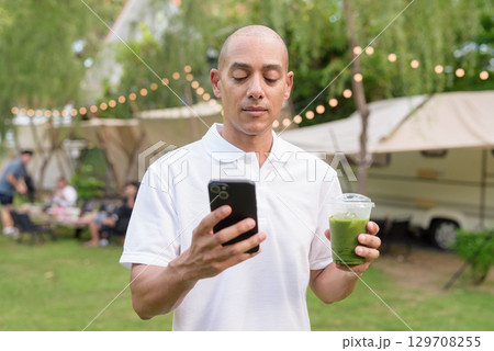 Bald man at campsite with camper van having healthy green detox juice drink and using phone 129708255