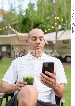 Bald man at campsite with camper van having healthy green detox juice drink and using phone 129708309