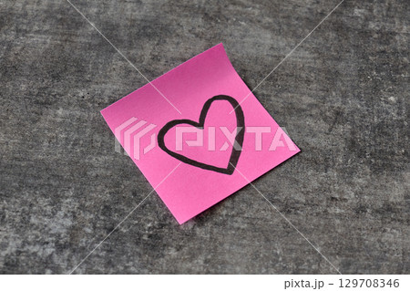 A pink sticky note with a heart sketch rests on a gray surface A pink sticky note with a heart sketch rests on a gray surface 129708346