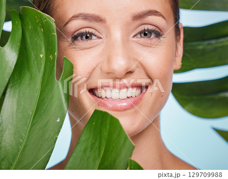 Portrait, skincare and palm leaf with a model woman posing in studio on blue background for beauty. Face, skin or nature with an attractive young female standing behind a plant for natural treatment Portrait, skincare and palm leaf with a model woman posing in studio on blue background for beauty. Face, skin or nature with an attractive young female standing behind a plant for natural treatment 129709988