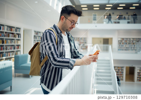 Smartphone, library and student on university, education or college website, internet search and school learning faq, about us or contact. Campus, bookshelf and geek man using phone for social media 129710308