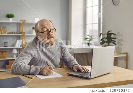 Senior man in eyeglasses using laptop and making notes on paper 129710375