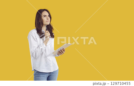 Woman standing on yellow copy space background, holding clipboard and thinking what to write Woman standing on yellow copy space background, holding clipboard and thinking what to write 129710455