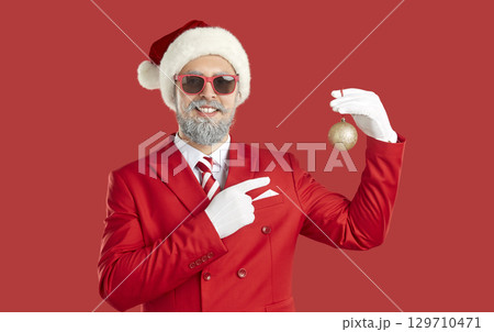 Man in red suit, hat and sunglasses standing on red background and holding Christmas ball 129710471