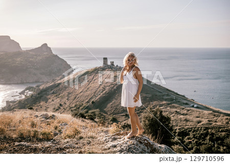 A blonde woman stands on a hill overlooking the ocean. She is wearing a white dress and she is enjoying the view. 129710596