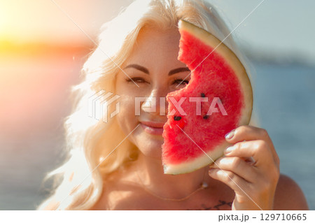 Woman Watermelon Beach Summer: Outdoors portrait, blonde woman playfully holding a bitten watermelon slice near sea, sunny day. Woman Watermelon Beach Summer: Outdoors portrait, blonde woman playfully holding a bitten watermelon slice near sea, sunny day. 129710665