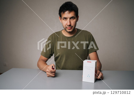 Ufa, Russia - 21 July 2025. Portrait of influencer male unpacking new IQOC tobacco heating system, holding heated tobacco unit, sitting at table on isolated background, looking at camera. 129710812