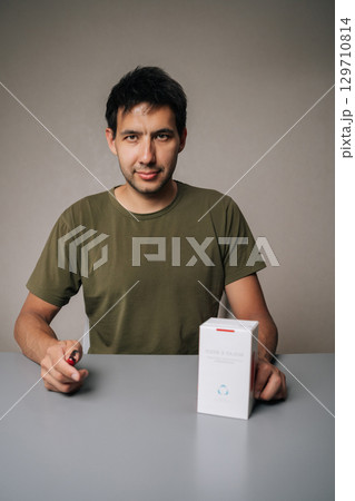 Ufa, Russia - 21 July 2025. Vertical portrait of handsome young man unpacking new IQOC tobacco heating system, holding heated tobacco unit, sitting at table on isolated background, looking at camera. Ufa, Russia - 21 July 2025. Vertical portrait of handsome young man unpacking new IQOC tobacco heating system, holding heated tobacco unit, sitting at table on isolated background, looking at camera. 129710814