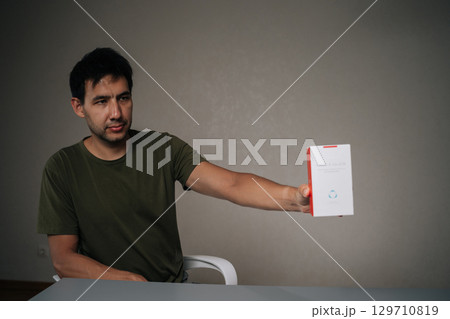 Ufa, Russia - 21 July 2025. Handsome young man unpacking new IQOC tobacco heating system, holding heated tobacco unit in hands , sitting at table on isolated background, looking away. 129710819