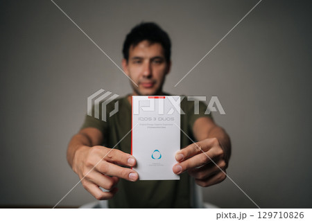 Ufa, Russia - 21 July 2025. Blurred portrait of influencer male holding and presenting new IQOC 3 duos tobacco heating system, innovative electronic cigarette for modern smokers seeking harm reduction 129710826