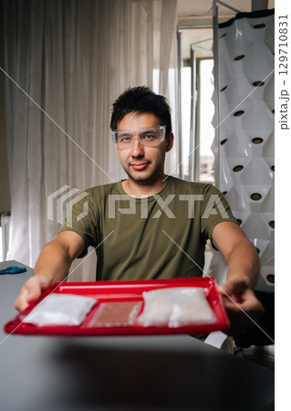 Vertical portrait of plant researcher in safety glasses holds tray of various fertilizers, preparing for planting in vertical hydroponic tower, highlighting nutrient solutions in modern agriculture. Vertical portrait of plant researcher in safety glasses holds tray of various fertilizers, preparing for planting in vertical hydroponic tower, highlighting nutrient solutions in modern agriculture. 129710831
