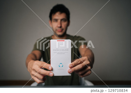 Ufa, Russia - 21 July 2025. Caucasian man holding and presenting new IQOC 3 duos tobacco heating system, innovative electronic cigarette alternative designed for modern smokers seeking harm reduction 129710834