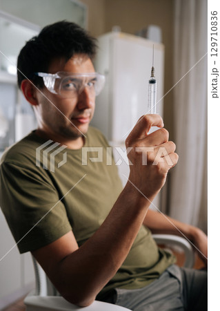 Vertical portrait of chemist wearing safety glasses carefully preparing fertilizer using syringe, ensuring precise measurements for optimal plant growth and healthy harvest, looking at camera. Vertical portrait of chemist wearing safety glasses carefully preparing fertilizer using syringe, ensuring precise measurements for optimal plant growth and healthy harvest, looking at camera. 129710836