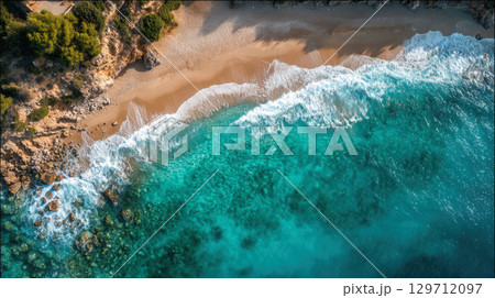 An exquisite aerial perspective captures the pristine beauty of a secluded beach, where vibrant turquoise waters gently caress the golden sand alongside dramatic rocky cliffs and lush green foliage. 129712097