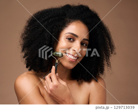 Face, skincare and portrait of black woman with roller in studio on a brown background. Makeup, cosmetics and happy female model with jade stone product for facial massage, beauty and healthy skin. 129713039