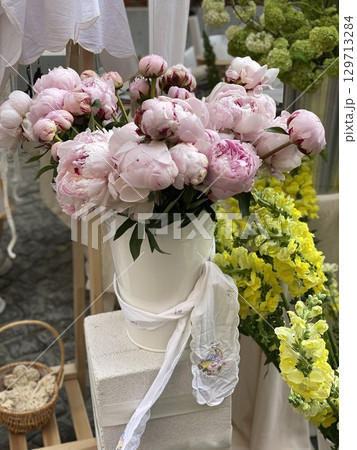 Bouquet of pink peonies in a white vase surrounded by yellow flowers at an outdoor market Bouquet of pink peonies in a white vase surrounded by yellow flowers at an outdoor market 129713284