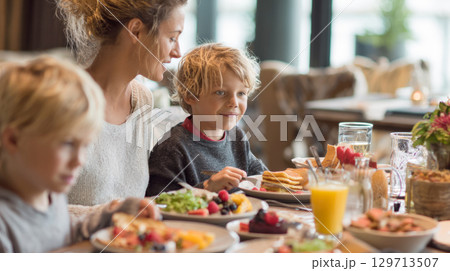 family sharing a delightful brunch. The mother smiles as her children enjoy their food, surrounded by plates of pancakes, fruit, and drinks, capturing a moment of togetherness and happiness 129713507